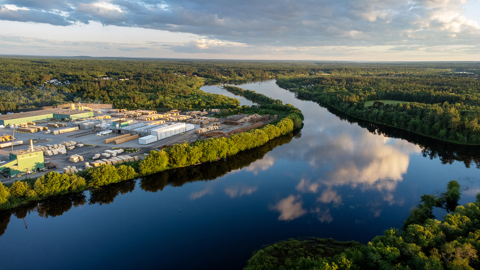 Aerial view of sawmill and timber kilns by a river, surrounded by dense forest and landscape.