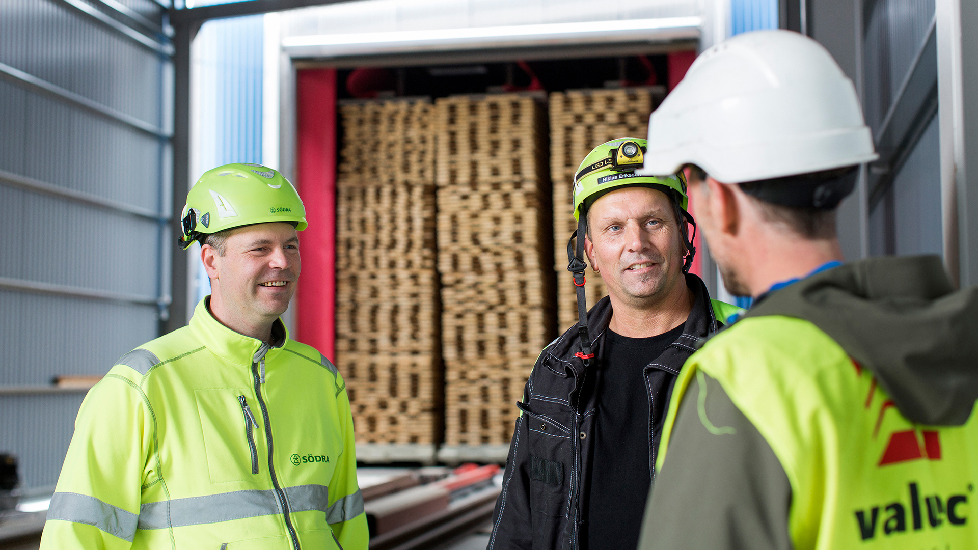 Three people in safety helmets talking in front of stacked timber inside a batch kiln.